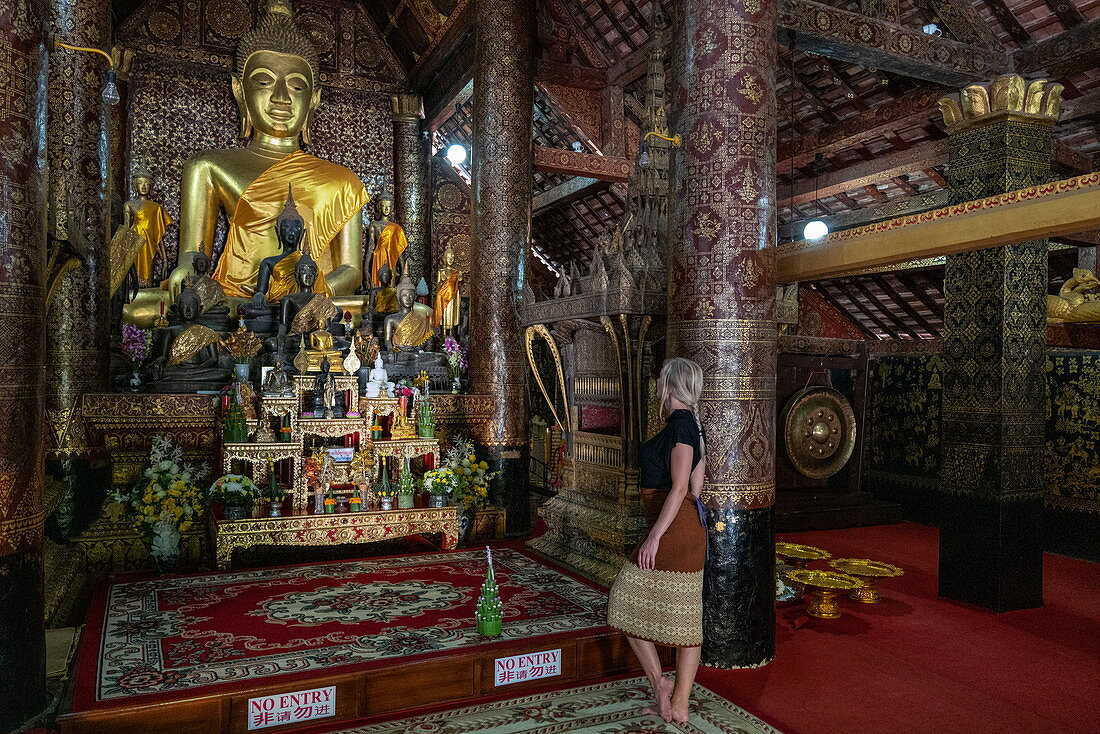 Junge blonde Frau bewundert die Buddha-Statue im buddhistischen Tempel Wat Xieng Thong (Tempel der goldenen Stadt), Luang Prabang, Provinz Luang Prabang, Laos, Asien