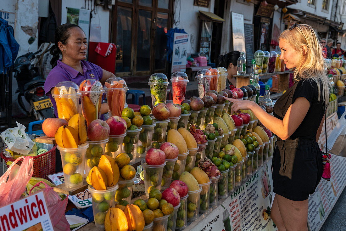 Junge blonde Frau an einem Marktstand an der Sisavangvong Road (der Hauptstraße) der frisch gepressten Fruchtsaft verkauft, Luang Prabang, Provinz Luang Prabang, Laos, Asien