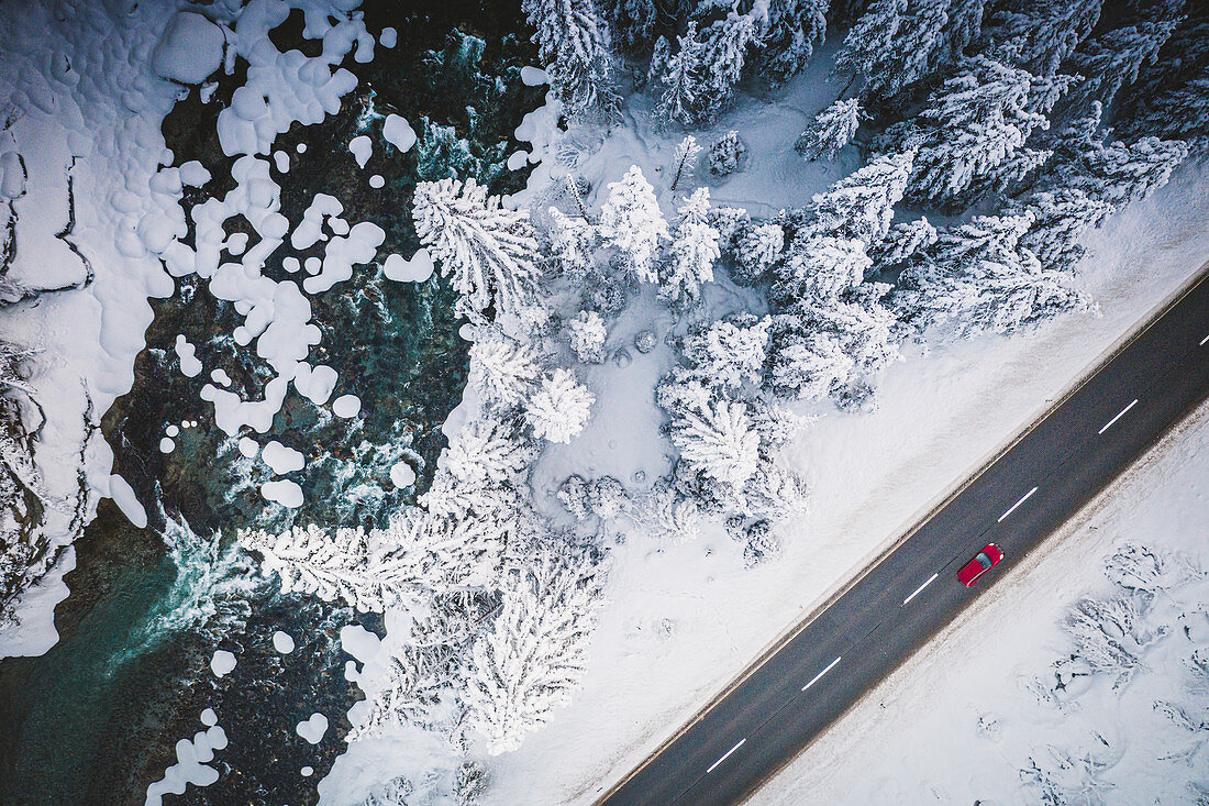 Auto, das auf der schneebedeckten Bergstraße auf Seite des gefrorenen Flusses und des Waldes, Luftbild, Schweiz, Europa reist