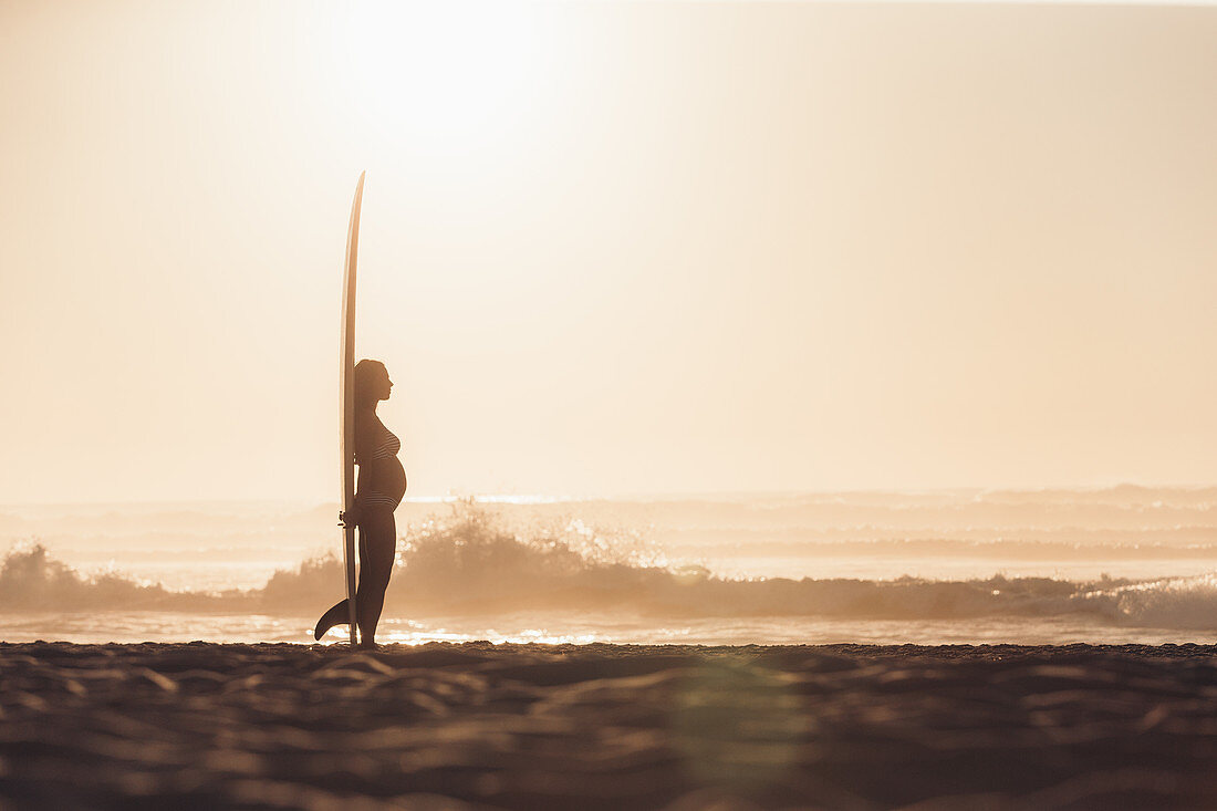 Schwangere Frau steht mit Surfbrett am Strand, Portugal