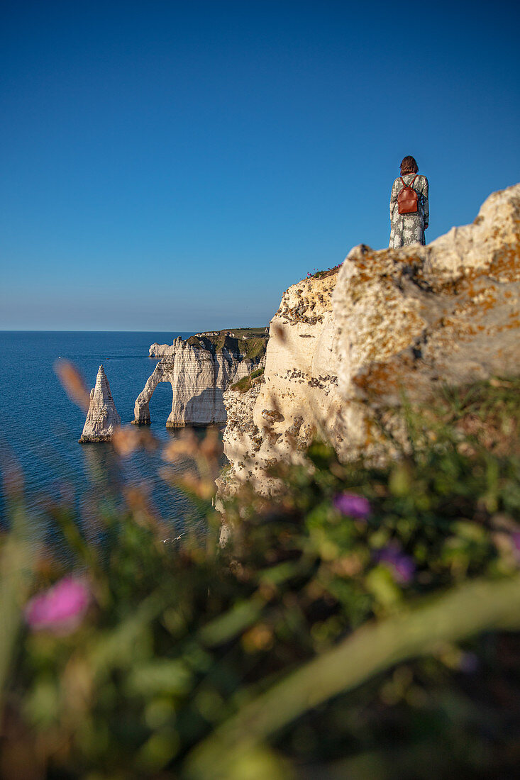 Eine Frau steht an der Alabasterküste und betrachtet den Felsbogen Porte d'Aval (Elefantenrüssel) und die Felsnadel Aiuille bei Étretat, Normandie, Frankreich