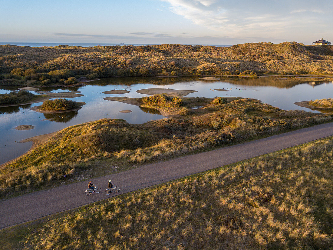 Luftaufnahme von zwei Menschen, die bei Sonnenuntergang auf einer Straße vorbei an Seen und Dünen entlang der Nordseeküste radeln, Midsland aan Zee, Terschelling, Westfriesische Inseln, Friesland, Niederlande, Europa