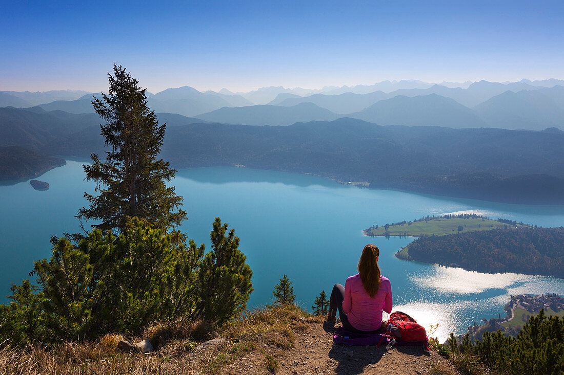 Woman takes a break from hiking, view from Herzogstand over Walchensee to the mountain range of the Alps, Bavaria, Germany