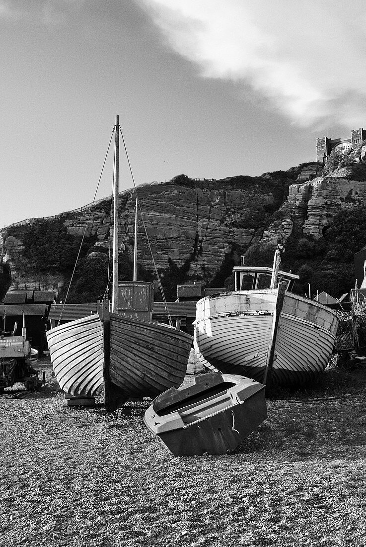 Traditionelle Fischerboote aus Holz auf dem Kiesstrand von Rock A Nore Beach mit Sandsteinfelsen und Standseilbahn im Hintergrund, Hastings, East Sussex, UK