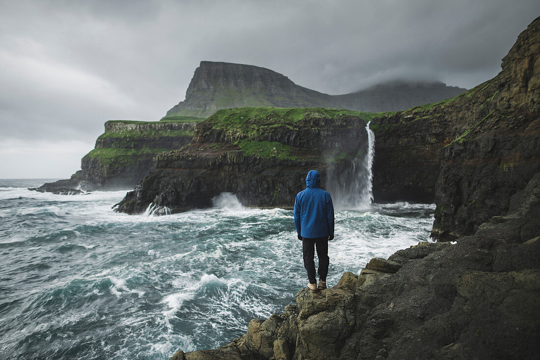 Denmark,Faroe Islands,Gasadalur Village,M?Lafossur Waterfall,Man standing on cliff and looking at Mulafossur Waterfall day during storm