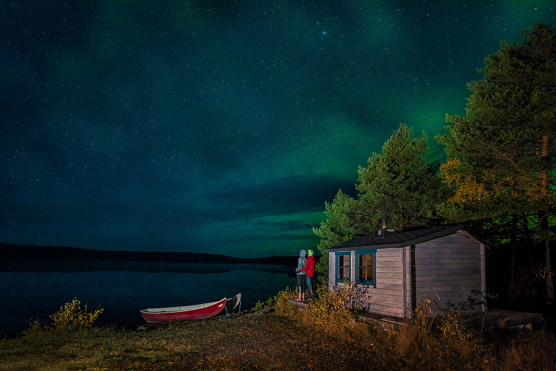 Couple watching polar lights in night sky at lakeside cabin with boats in Lapland, Sweden