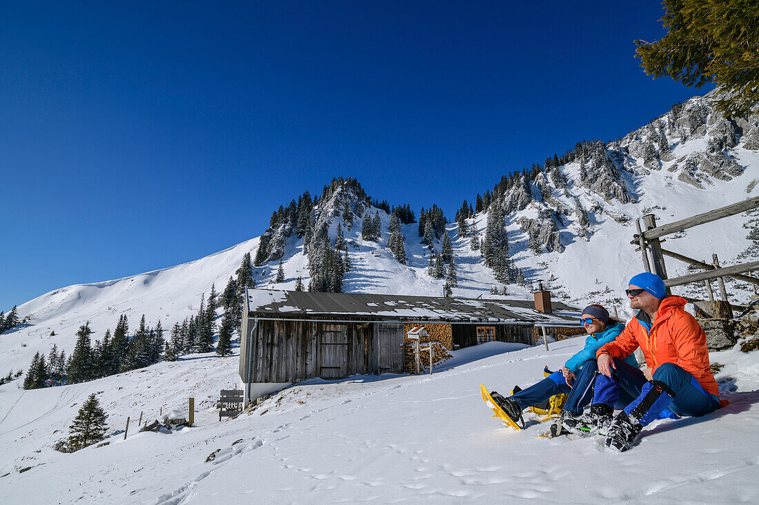 Mann und Frau beim Wandern sitzen im Schnee vor Hütte, Obere Schönfeldalm, Jägerkamp, Spitzinggebiet, Bayerische Alpen, Oberbayern, Bayern, Deutschland