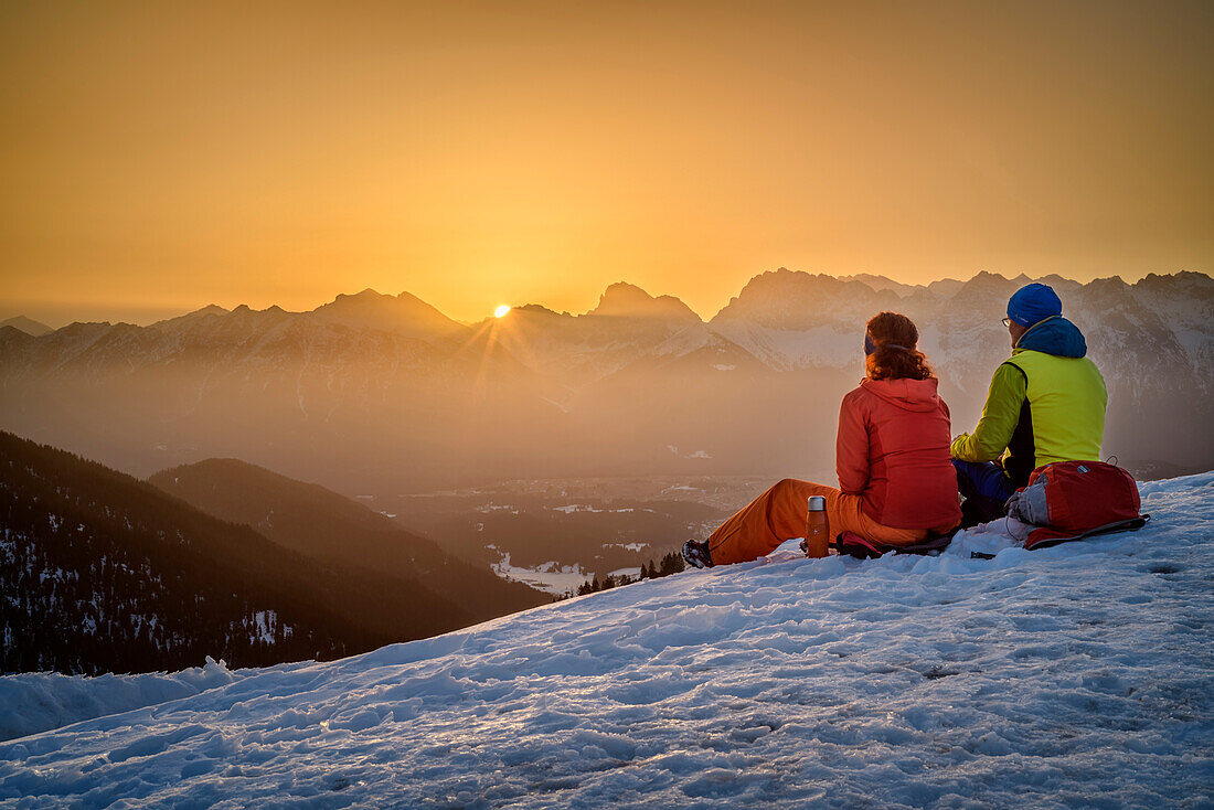 Mann und Frau beim Wandern sitzen im Schnee und blicken in Sonnenaufgang, Wank, Estergebirge, Bayerische Alpen, Oberbayern, Bayern, Deutschland