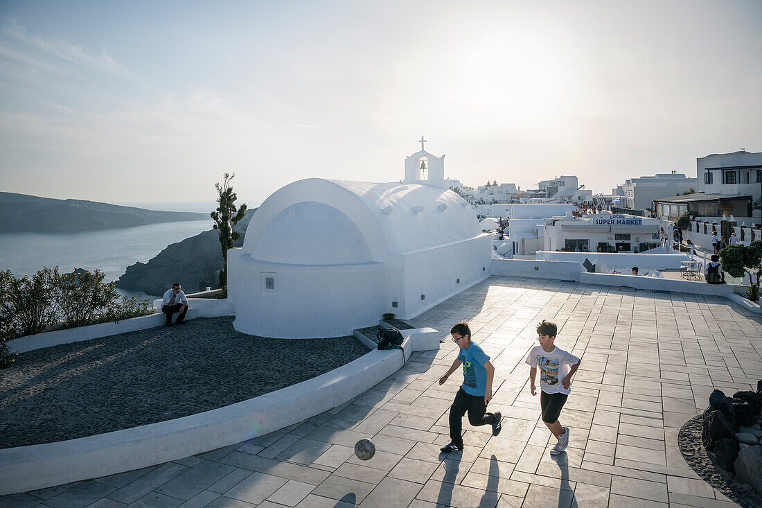 Kinder spielen Fussball am Vorplatz zur Kapelle mit Blick auf Caldera, Oia, Santorini, Santorin, Kykladen, Ägäisches Meer, Mittelmeer, Griechenland, Europa