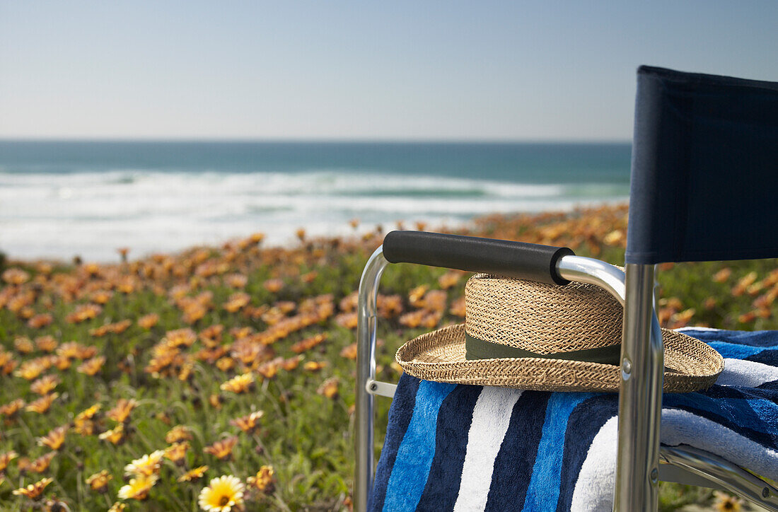 Sonnenhut und Handtuch aus Stroh auf einem leeren Regiestuhl, der auf einer Bank voller Blumen mit Blick auf den Strand sitzt