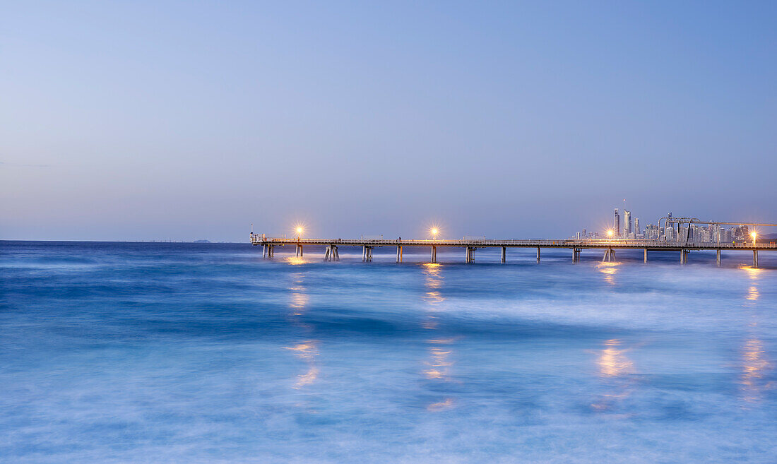 Long pier stretching out into calm ocean in early evening with lights on