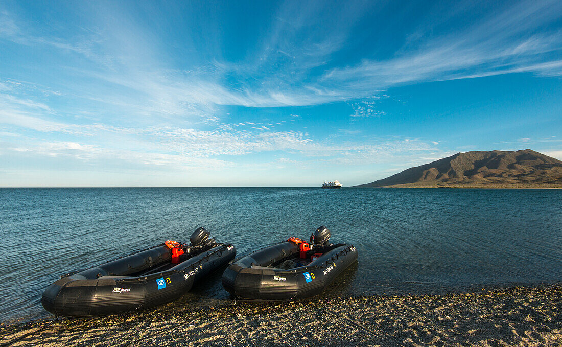 Zwei Zodiacs von Lindblad Expeditions warten bei Sonnenuntergang am Strand, während der National Geographic Sea Lion am Horizont vor Anker liegt. Magdalena-Insel, Baja California Sur.