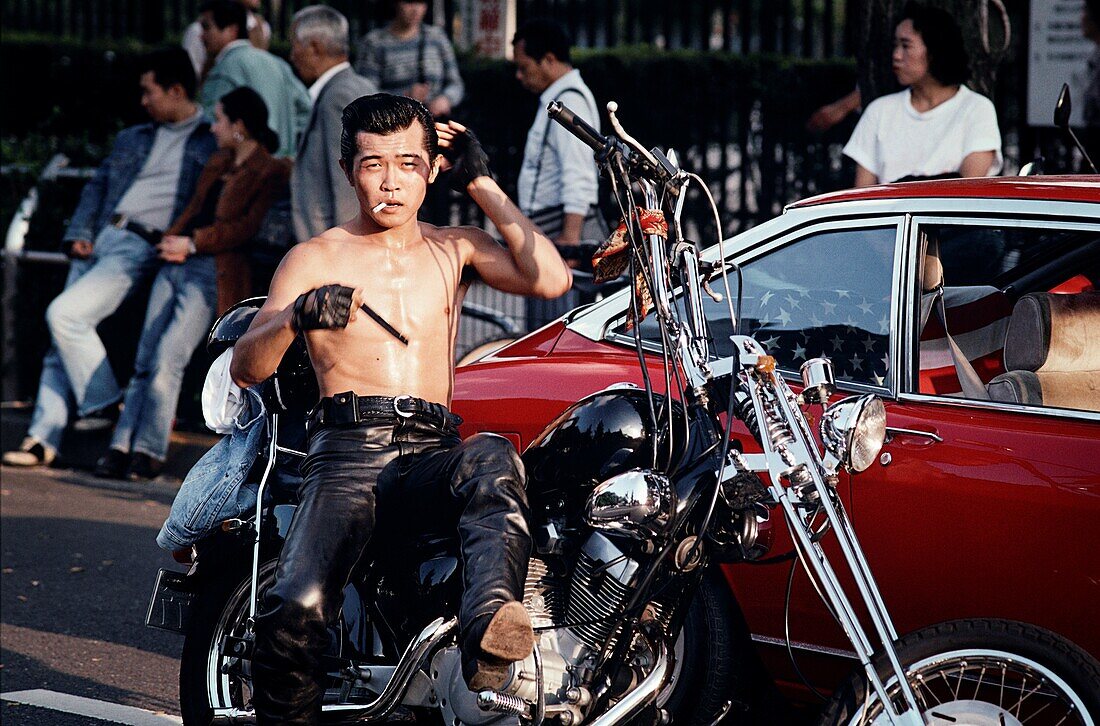 Hemdloser Mann in Lederhose, der eine Zigarette raucht und sich die Haare kämmt, während er auf einem Motorrad in einer belebten Straße sitzt, Tokio, Japan