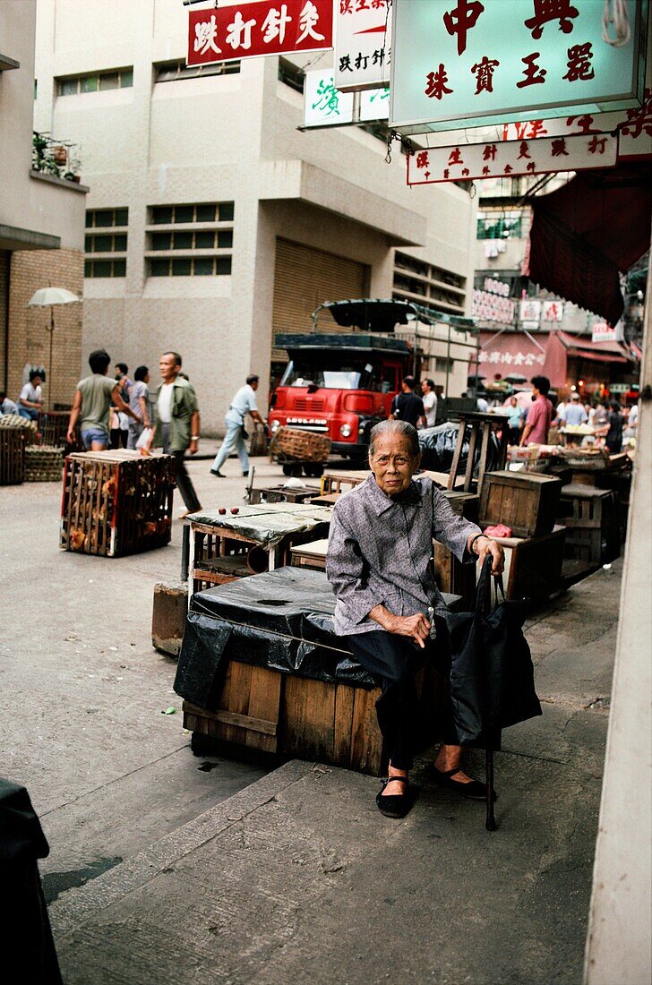 Frau mit Stock sitzt auf einer Holzkiste auf der Straße, Hongkong