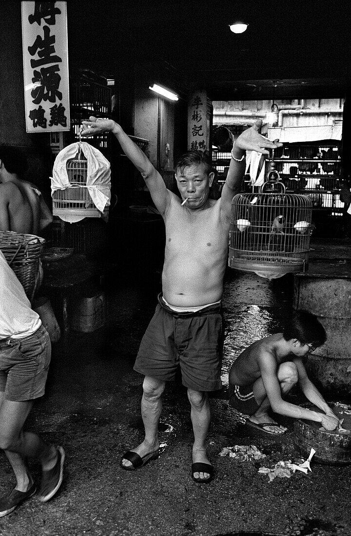 China, Hong Kong, Shirtless man with cigarette in his mouth holding up two birdcages at street market