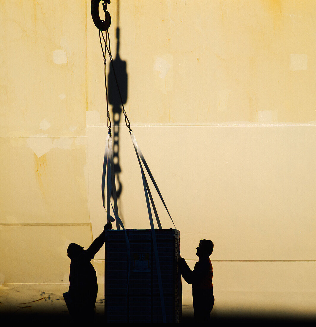 Silhouette von zwei Männern, die die Fracht führen, die an einem großen Haken vor dem Schiffsrumpf befestigt ist