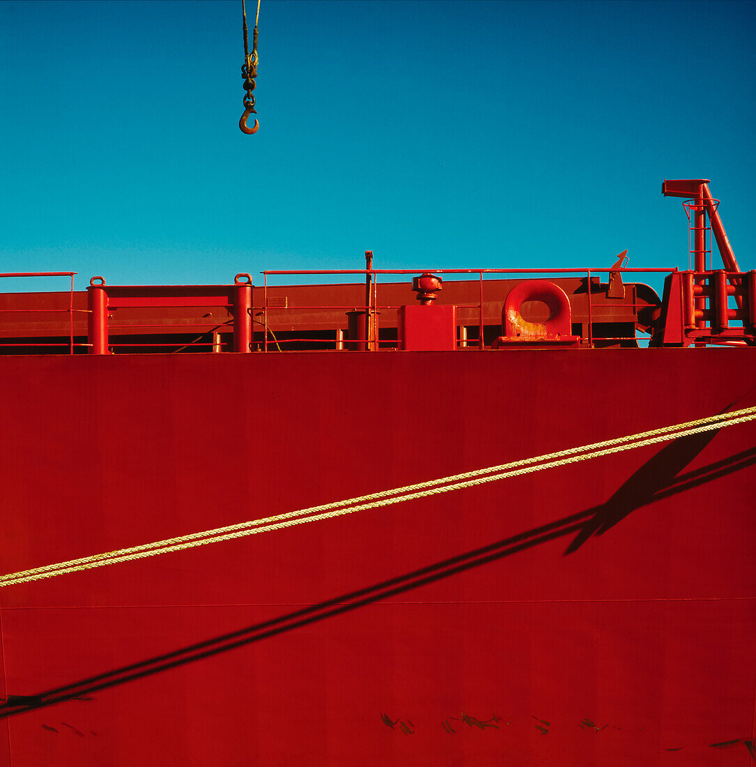 Kranhaken gegen den blauen Himmel, der zum roten Schiff absteigt, das an den Hafen gebunden ist