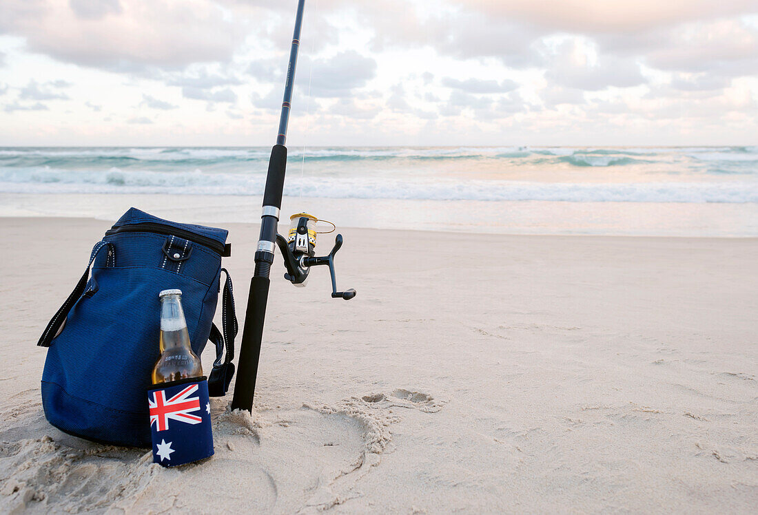 Bier im Stubbie-Halter mit australischer Flagge, Kühltasche und Angelrute im Sand am Strand