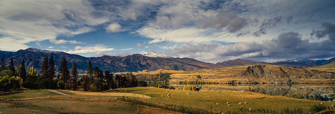Panorama of farm and snow capped mountain range surrounding Lake Hayes and round hay bales in field