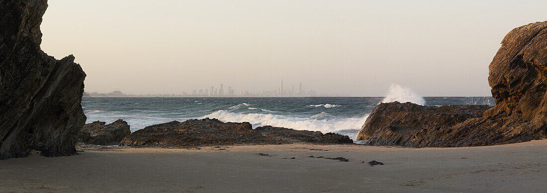 Blick zwischen großen Felsen mit brechenden Wellen - die Skyline von Surfers Paradise am Horizont