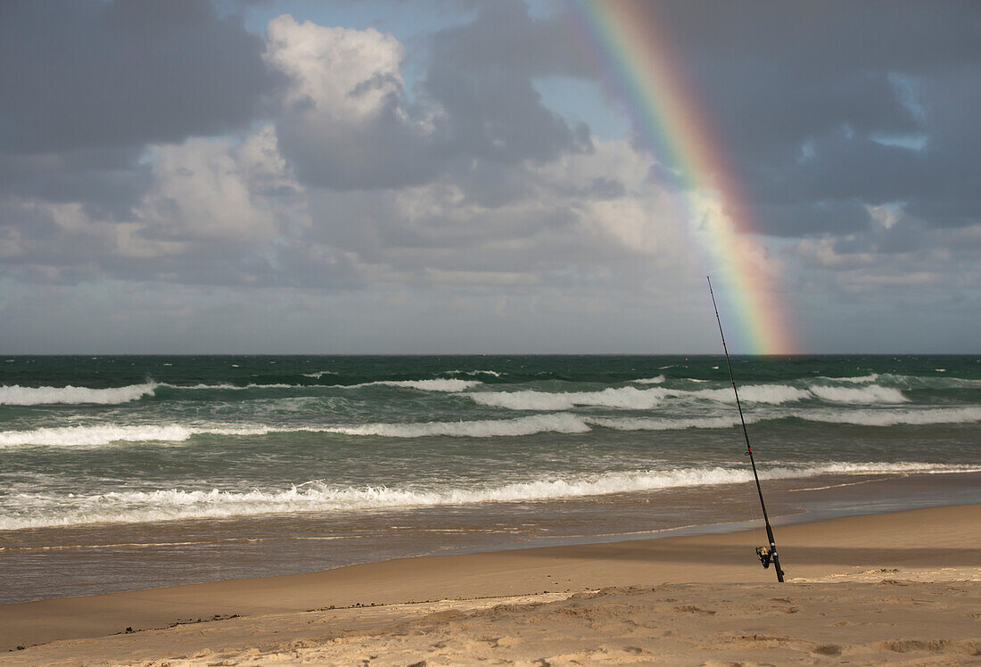 Surf Caster Angelrute im Sand und Regenbogen im stürmischen Himmel am Strand gesichert