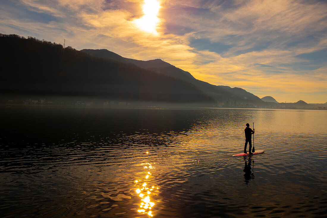 Mann beim SUP am Luganersee mit Sonnenlicht und Berg im Tessin, Schweiz