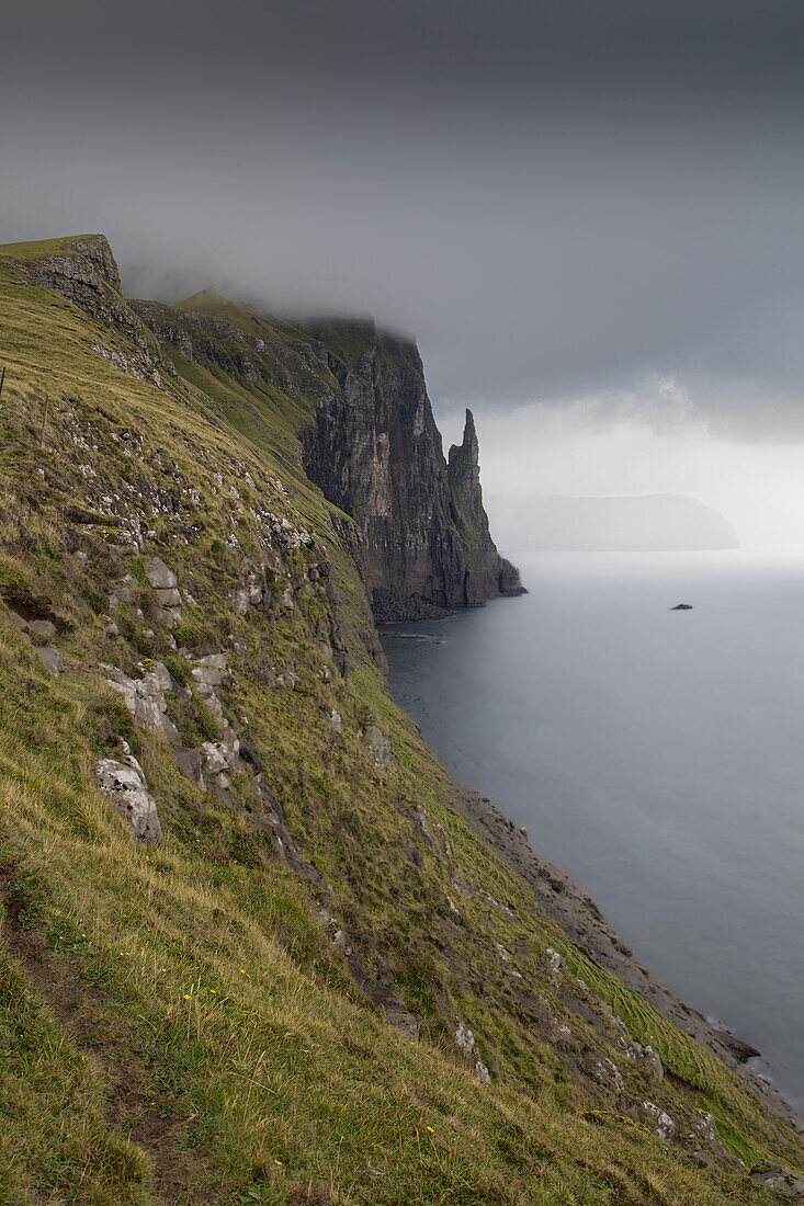 View along cliffs to Witches Finger, sea stack. Vagar, Faeroer. dark rain clouds.