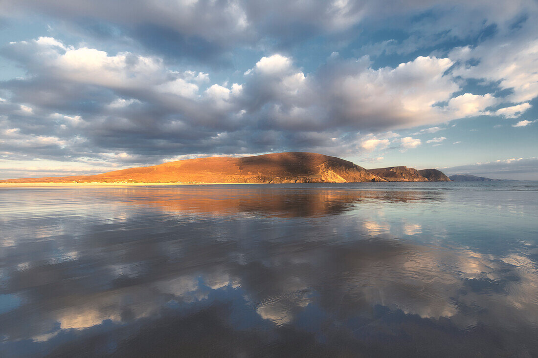 Range of hills and cloudy sky are reflected in the water on the evening beach. Keel Beach, County Mayo, Ireland.