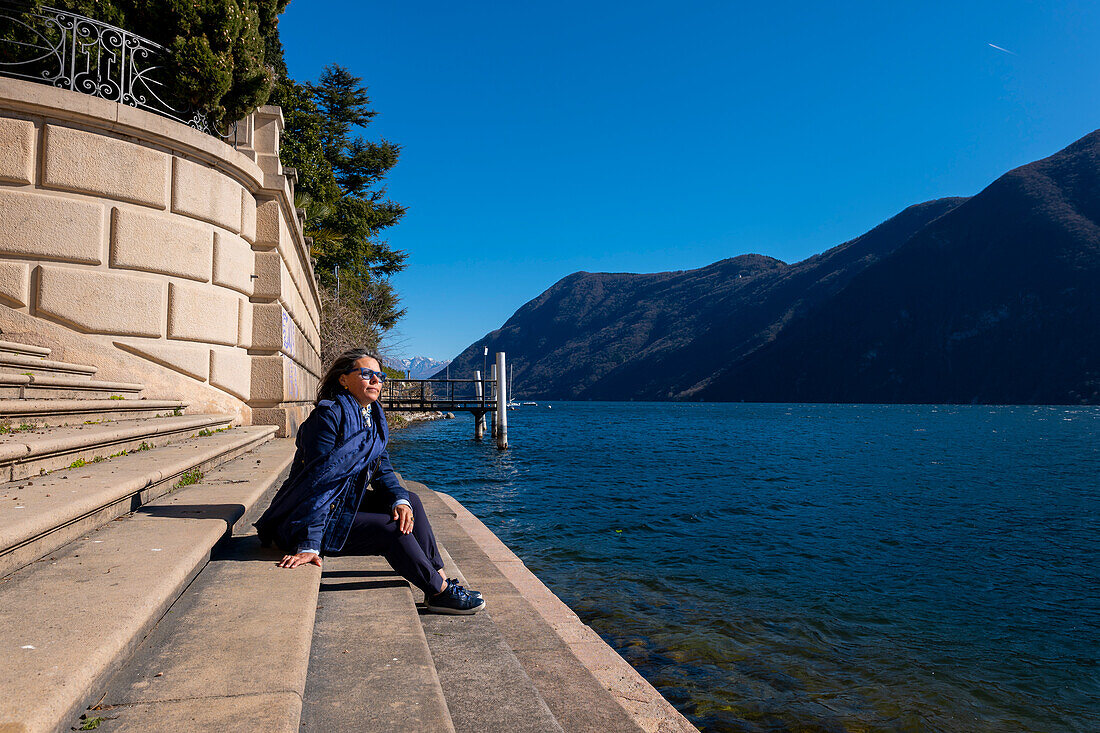 Frau sitzt auf einer Treppe am Wasser mit Blick auf die Berge über den Luganersee an einem sonnigen Tag in Lugano, Tessin, Schweiz.
