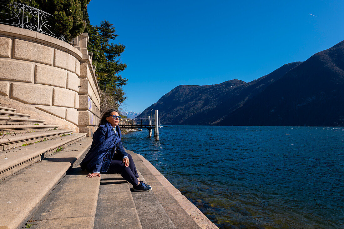 Woman Sitting on a Staircase on the Waterfront with Mountain View over Lake Lugano in a Sunny Day in Lugano, Ticino, Switzerland.