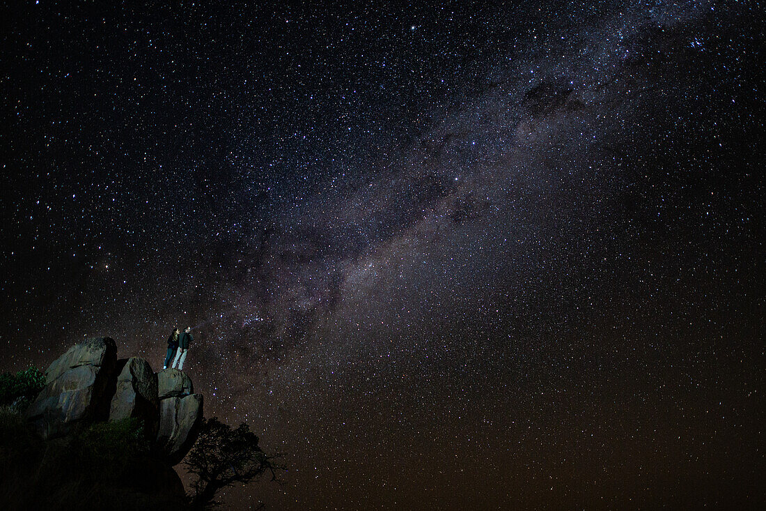 A man and woman stand on top of boulders looking up at the milk way