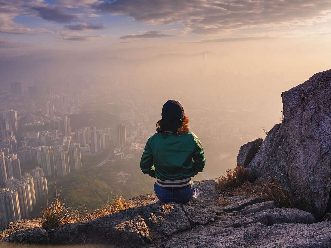 Frau sitzt auf Felsen in Hügeln über Hongkong und blickt über die Stadt