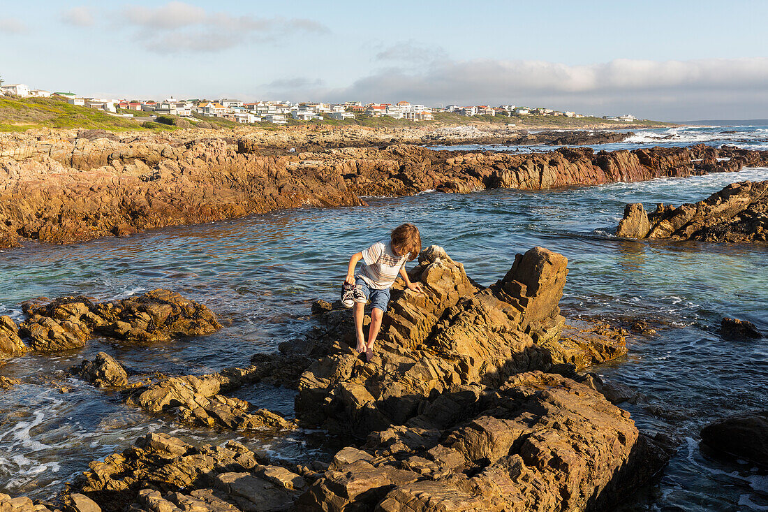 A young boy exploring the rock pools on a jagged rocky Atlantic Ocean coastline, De Kelders, Western Cape, South Africa.