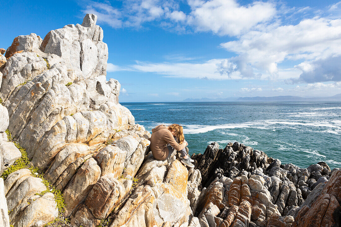 Teenager-Mädchen sitzt auf Felsen mit Blick auf den Atlantischen Ozean, ihr Kopf ruht auf ihren Knien