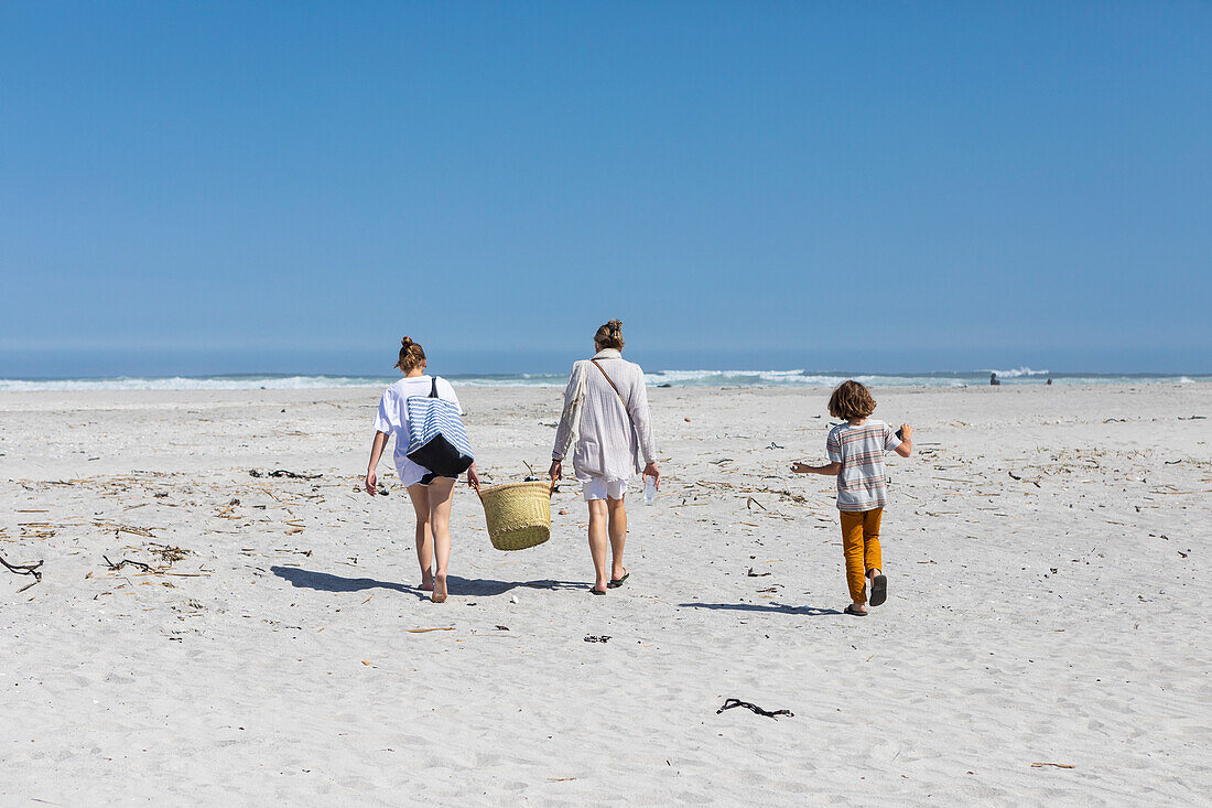 Mutter und Tochter im Teenageralter zu Fuß an einem Sandstrand mit einem Korb, Junge folgt, Grotto Beach, Südafrika