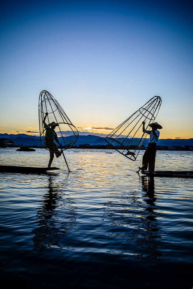 Two fishermen standing on their boats fishing on Lake Inle at dusk.
