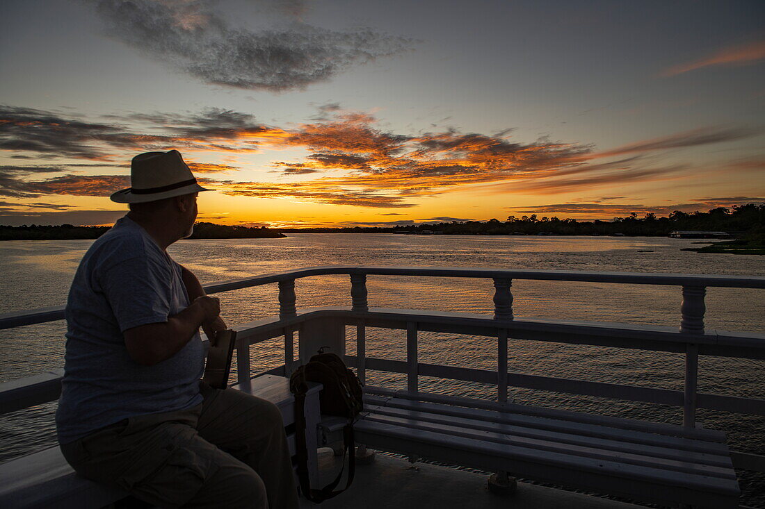 Silhouette von einem Mann der den Sonnenuntergang auf dem Außendeck eines kleinen Flussschiffes genießt, nahe Manaus, Amazonas, Brasilien, Südamerika