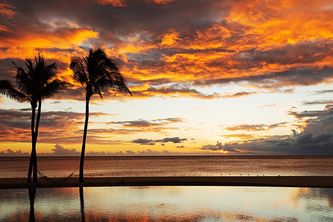 Palmen, die sich gegen rote Wolken abheben, spiegeln sich bei Sonnenuntergang über einem Strand in Flic en Flac, Mauritius, Indischer Ozean, Afrika in einem Infinity-Pool wider