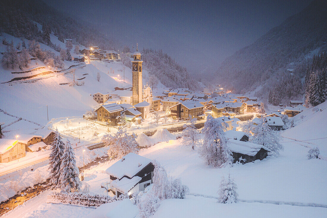 Beleuchtetes Dorf Gerola Alta und zugefrorener Fluss, der während der Winterdämmerung mit Schnee bedeckt ist, Valgerola, Veltlin, Lombardei, Italien, Europa
