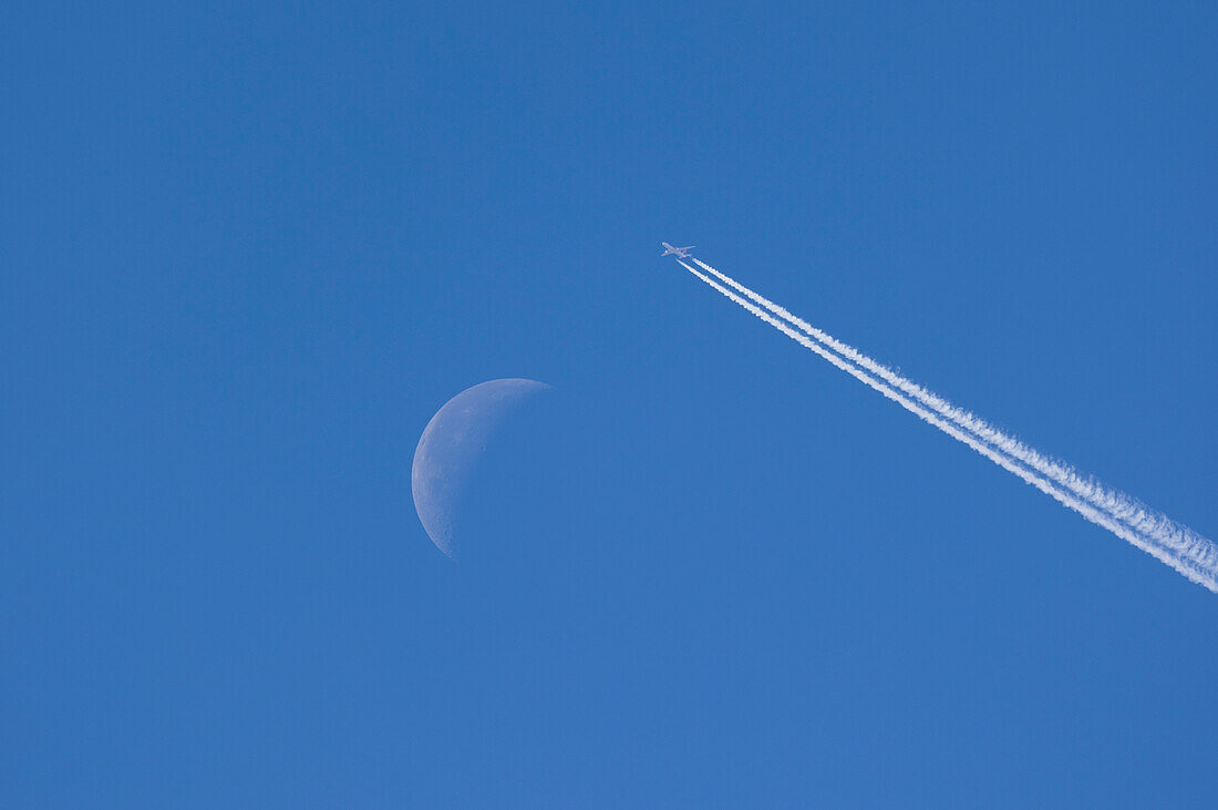 Flugzeug verlässt Kondensstreifen gegen blauen Himmel mit abnehmendem Mond