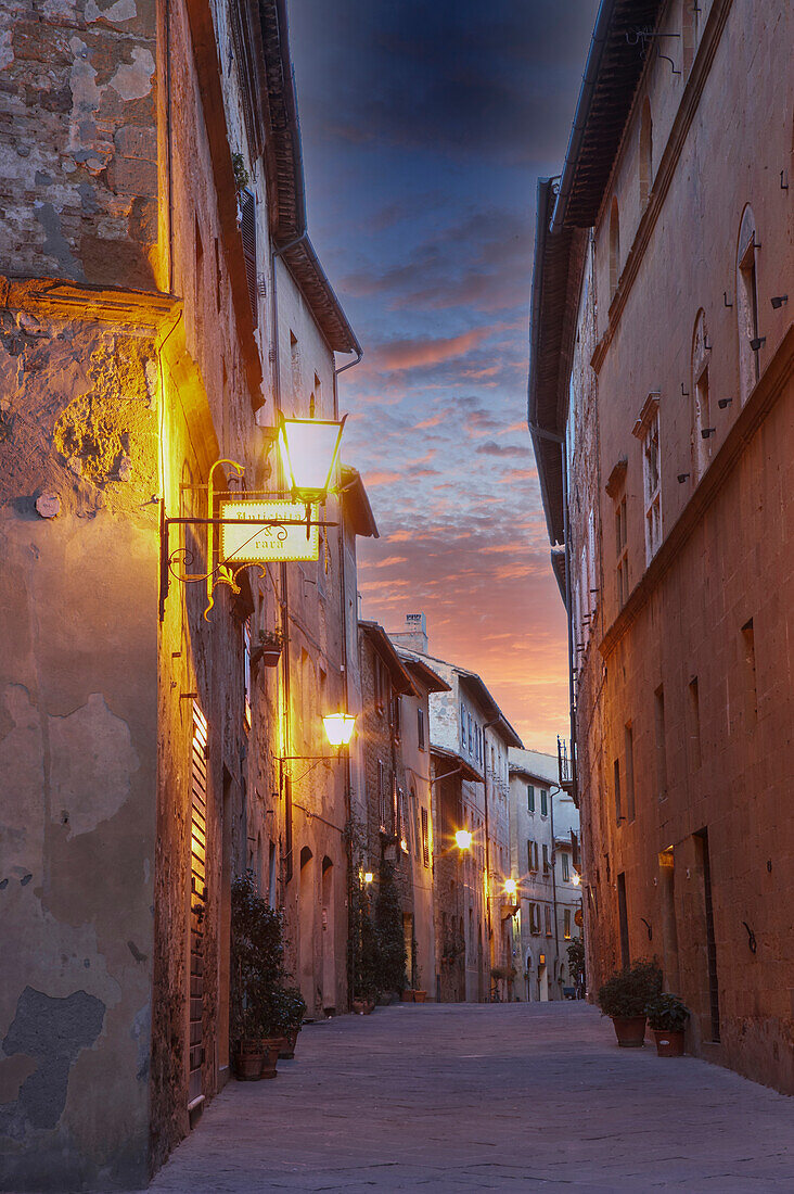 Italien, Toskana, Val D'Orcia, Pienza, schmale Gasse und beleuchtete Gebäude in der Altstadt in der Abenddämmerung