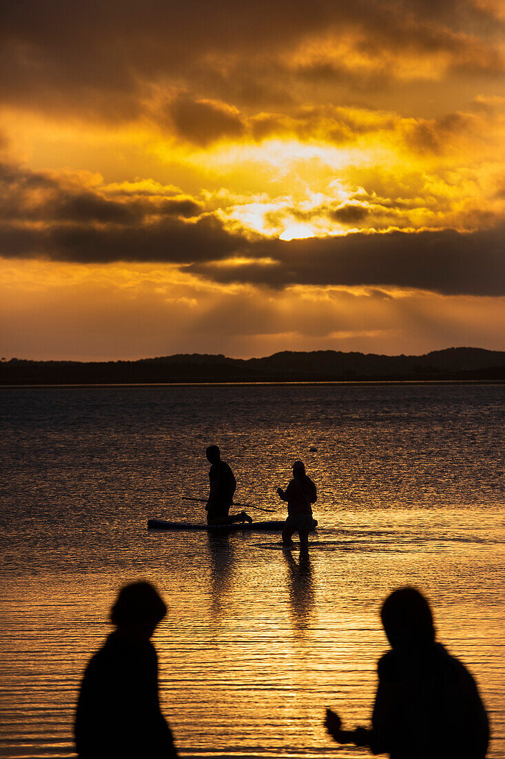 Silhouettierte Familie mit Paddleboard im Ozean unter dramatischem Himmel bei Sonnenuntergang