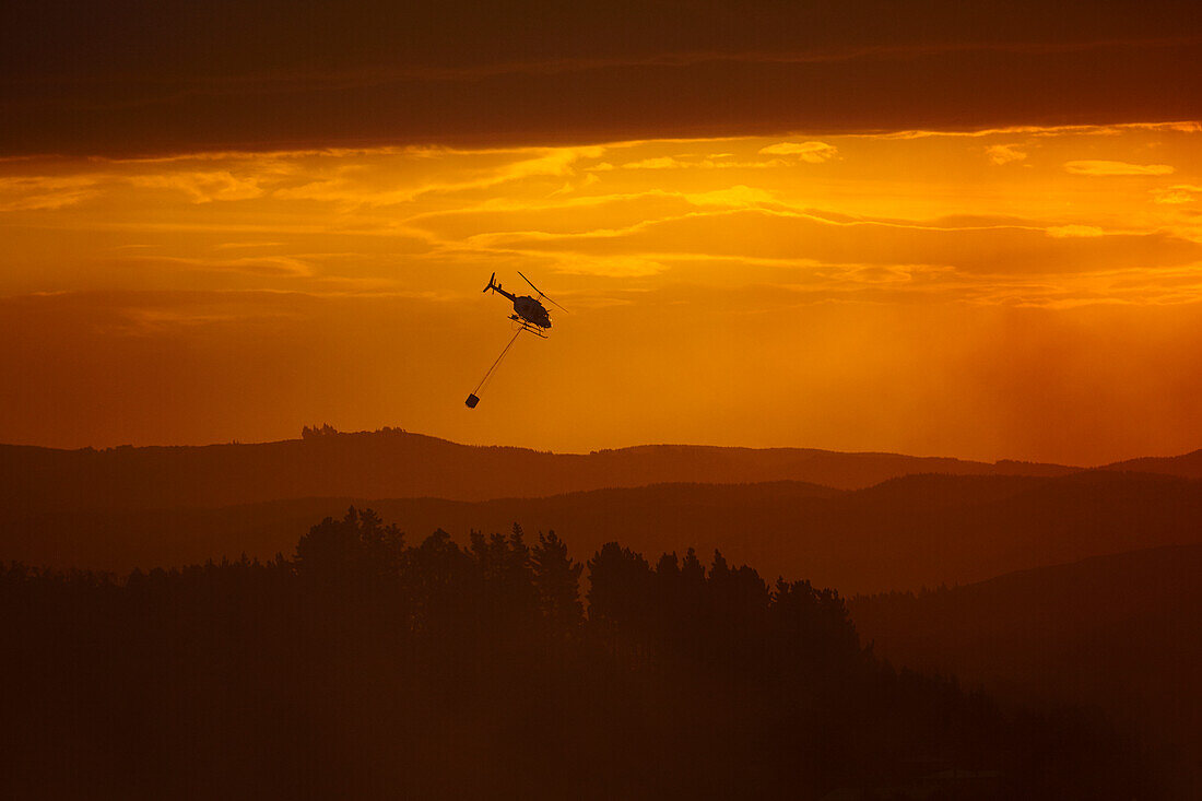 Rauchiger Sonnenuntergang und Hubschrauber bei der Brandbekämpfung in Burnside, Dunedin, Südinsel, Neuseeland