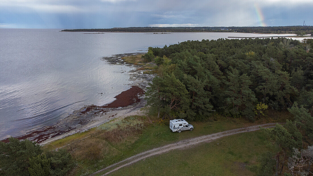 Einzelnes Wohnmobil steht an kleiner Straße am Meer, hinter Bäumen. Katthammarsvik, Gotland, Schweden.
