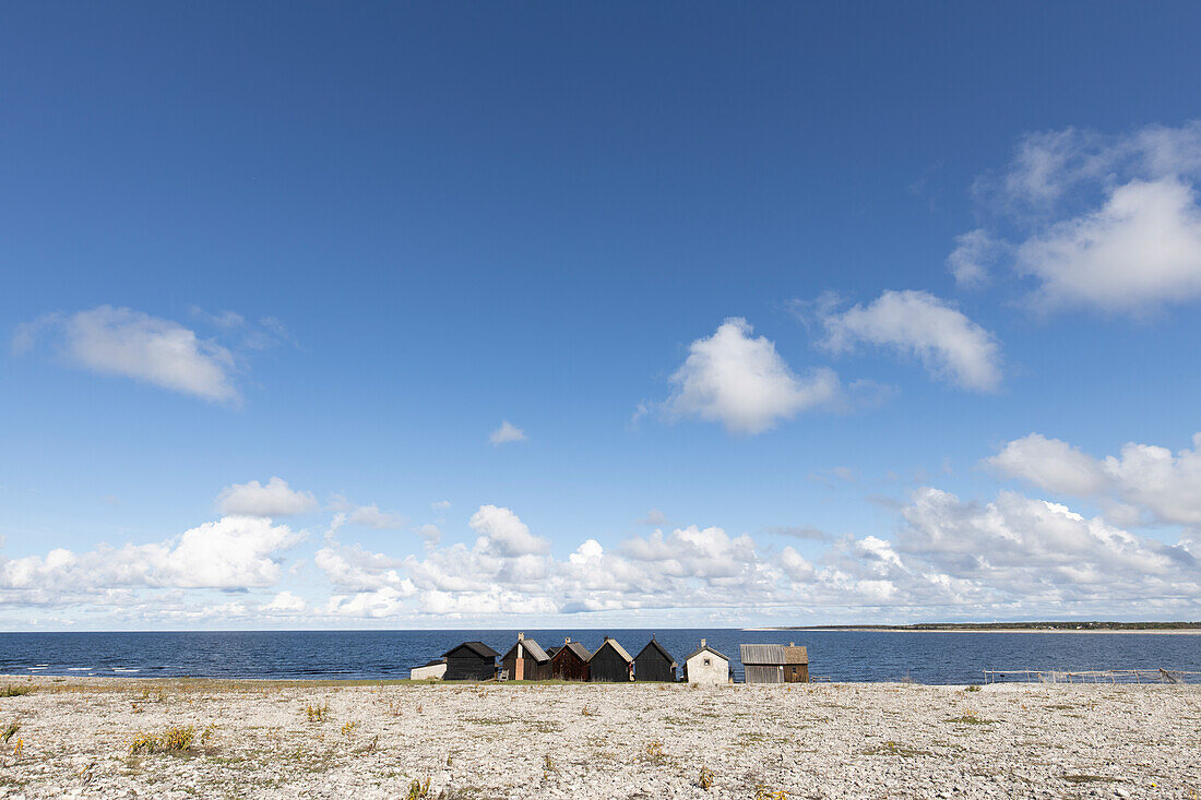 A row of lonely fisherman's huts stand on the stone beach. Fleecy clouds and blue sky. Helgumannen fiskelage. Faroe, Gotland County, Sweden.