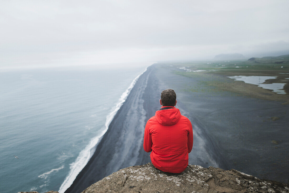 Mann mit rotem Mantel sitzt über dem Strand in Vik, Island