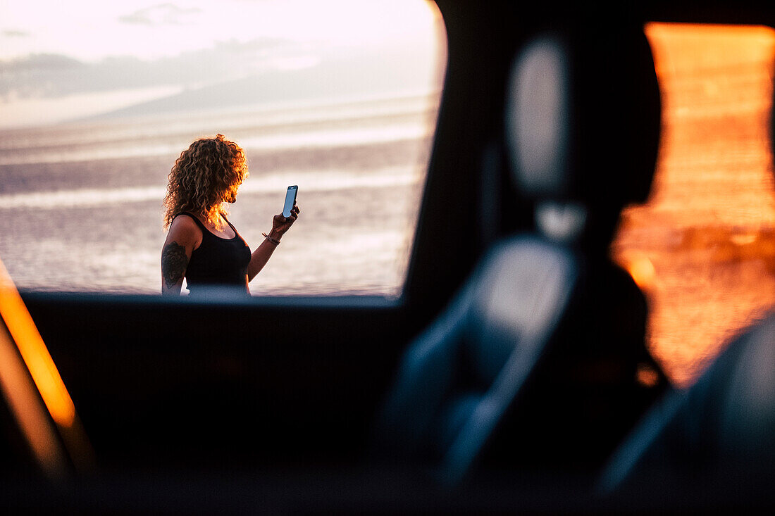 Frau mit Smartphone hinter Autofenster bei Sonnenuntergang