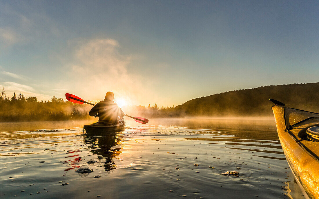 Kanada, British Columbia. Ein Kajakfahrer paddelt im sonnendurchfluteten frühen Morgennebel auf einem kanadischen See.