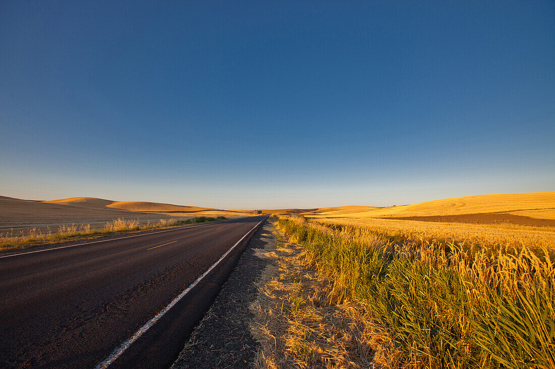 Eine Straße durch eine landwirtschaftliche Landschaft, gerade Straße und hügeliges Ackerland mit goldreifer Weizenernte.