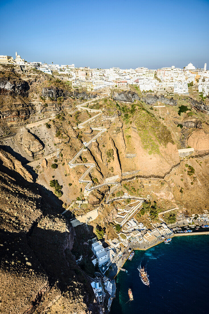 Erhöhter Blick auf eine steile Klippe, eine Stadt auf einem Hügel und einen Hafen auf dem Wasser mit einem Zickzackpfad die Klippe hinauf auf einer griechischen Insel.